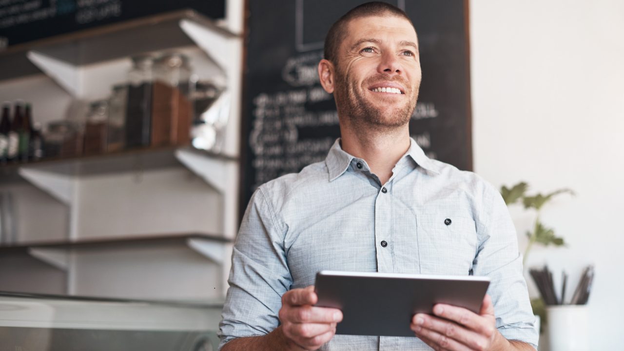 Smiling small business owner holding a tablet in his café, in front of a chalkboard menu and shelves of jars