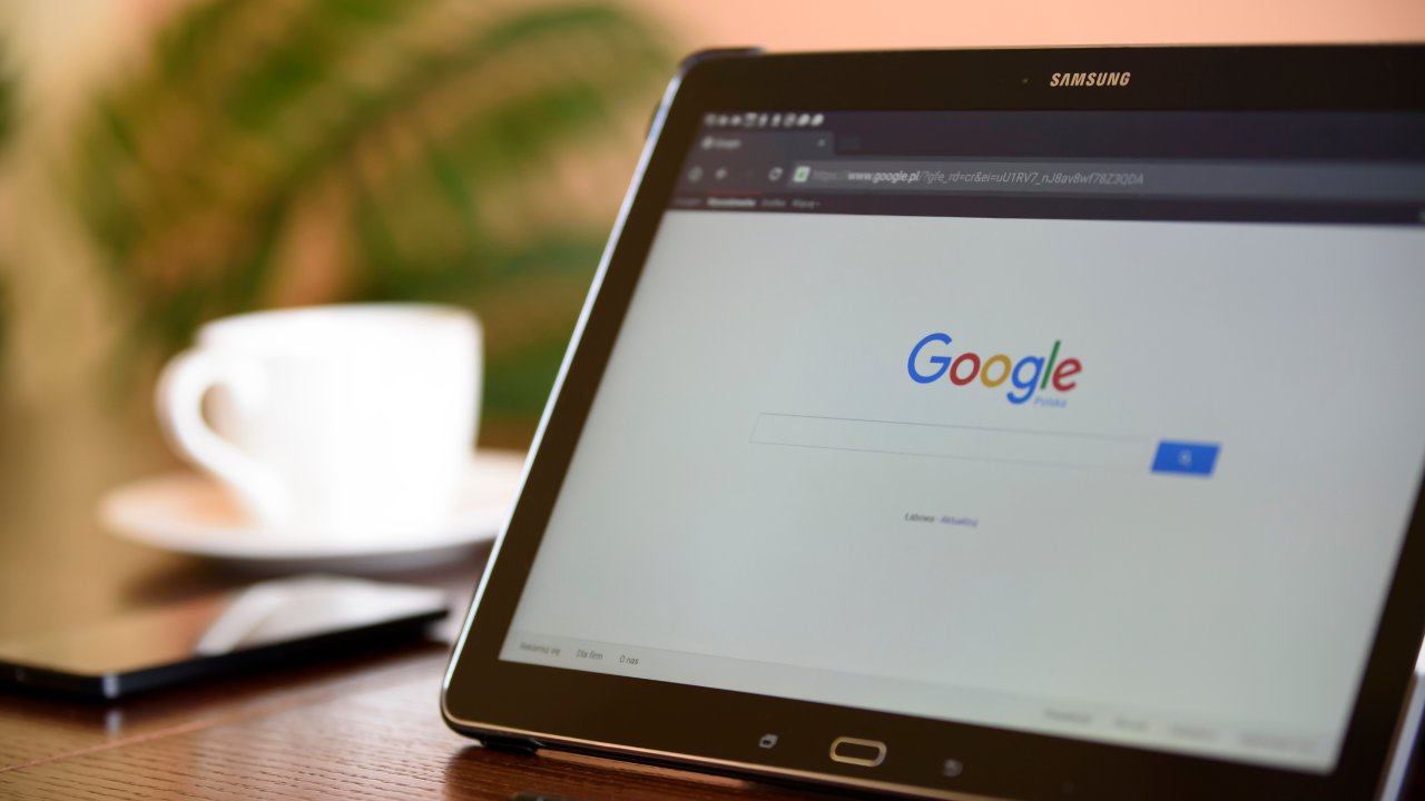 A tablet on a wooden desk showing a Google homepage, next to a coffee cup and a potted plant