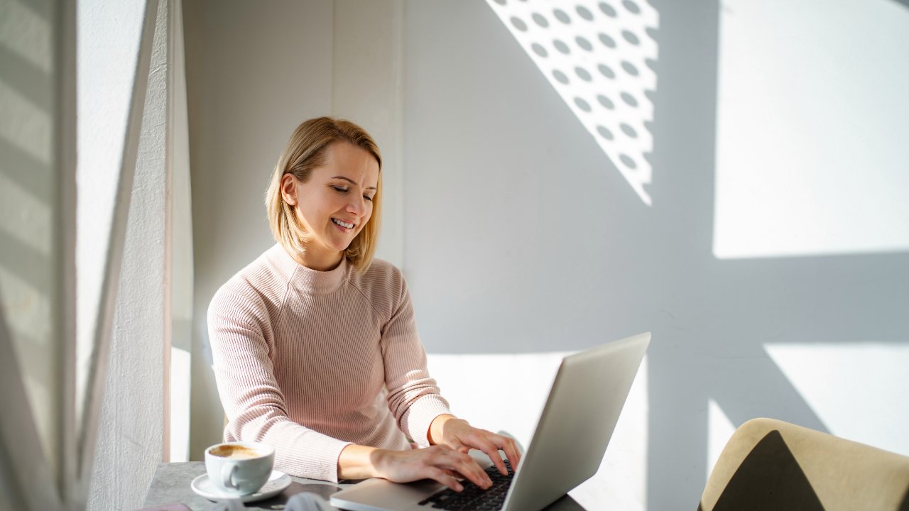 A woman smiling while working on a laptop in a sunlit office